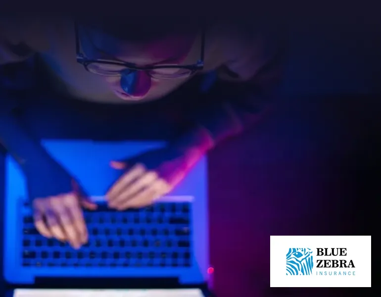 Person wearing glasses working intently on a laptop, their face illuminated by blue and purple light from the screen, with the Blue Zebra Insurance logo in the corner