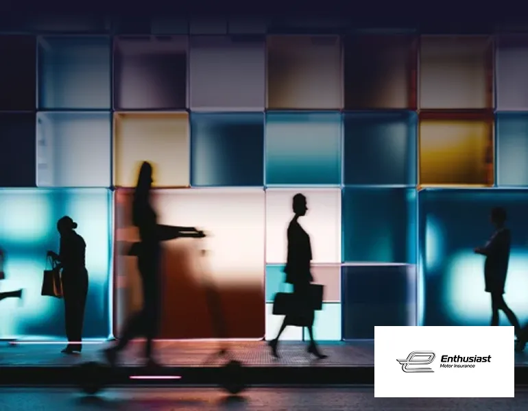 Silhouettes of business people walking past a modern building with colourful illuminated glass panels, captured with motion blur, with the Enthusiast Motor Insurance logo in the corner